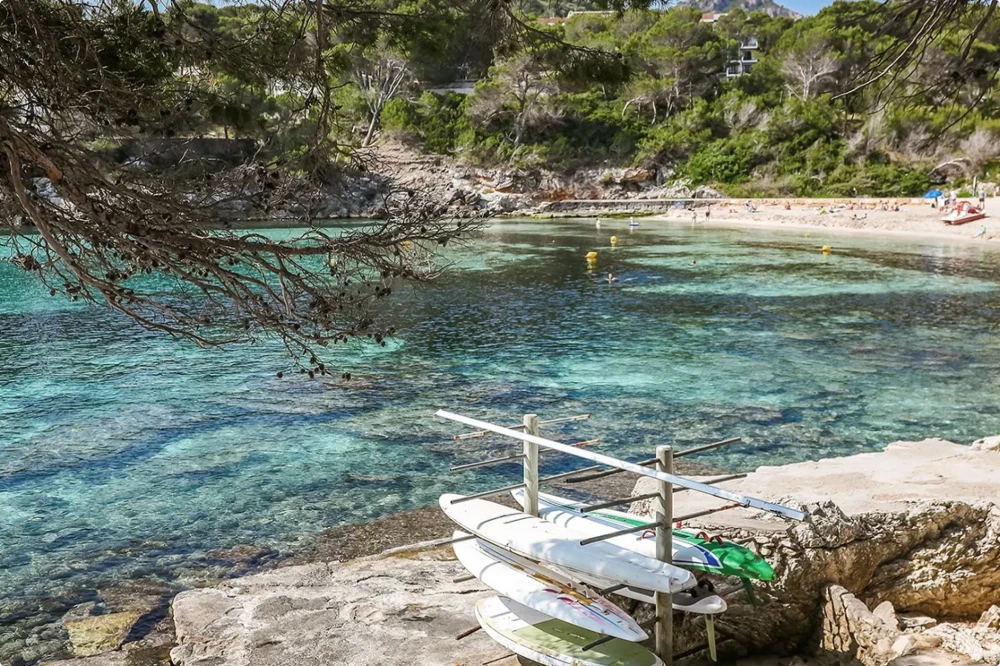 Tablas de paddle surf apiladas en una costa rocosa cerca de aguas turquesas cristalinas y una playa bañada por el sol.