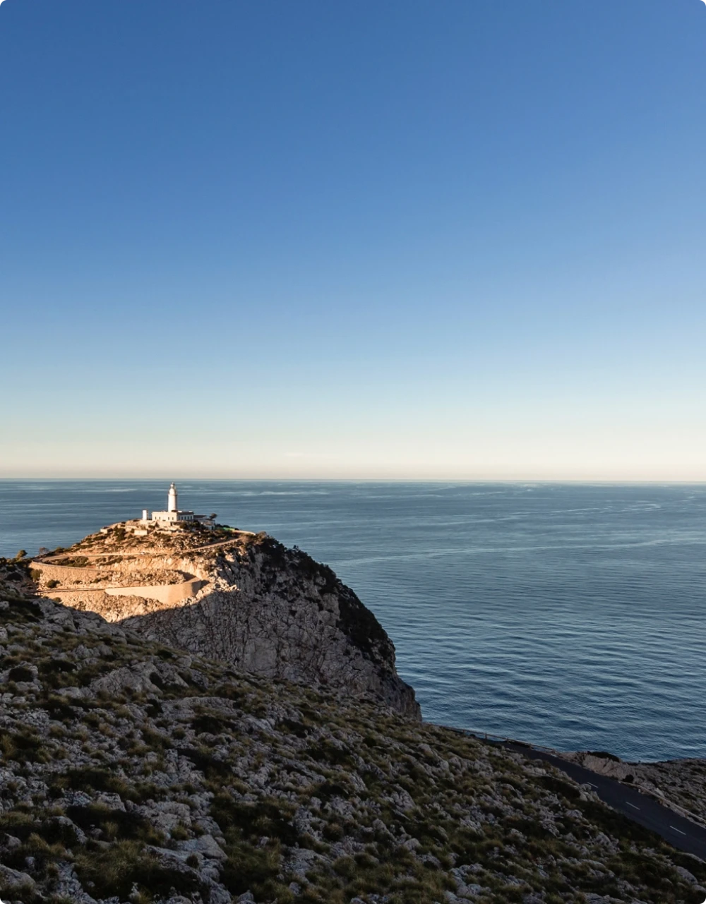 Un faro blanco encaramado en un acantilado rocoso y escarpado con vistas a un vasto y tranquilo mar azul bajo un cielo despejado.