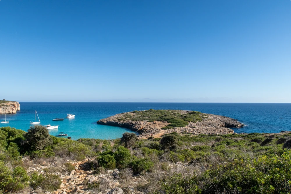 Paisaje costero con mar turquesa, una pequeña isla rocosa y barcos anclados en una bahía tranquila bajo un cielo azul claro.