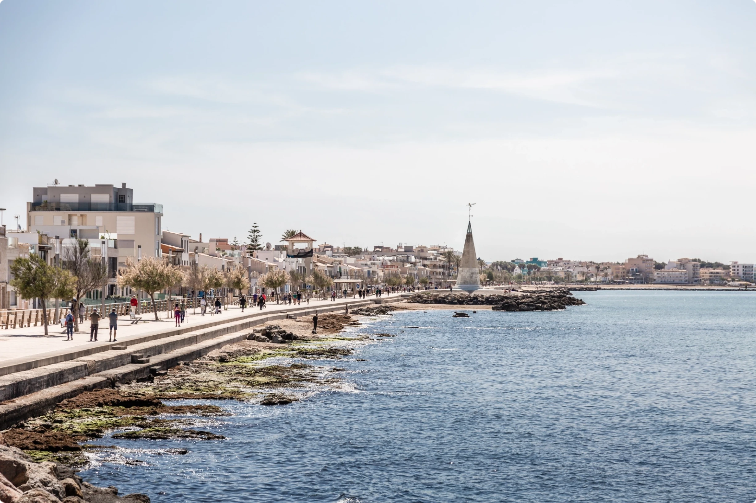 Paseo marítimo en Portixol, Mallorca, con gente paseando por la costa, edificios costeros y un mar azul tranquilo bajo un cielo despejado.