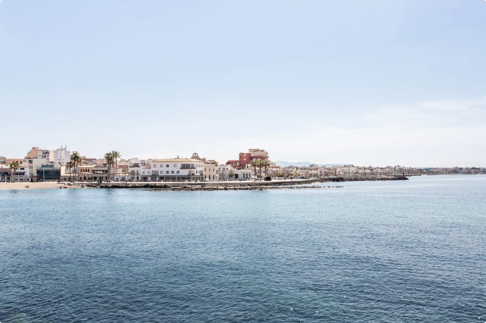 Vista costera de Portixol, Mallorca, con edificios frente al mar, palmeras y un mar azul tranquilo bajo un cielo despejado.