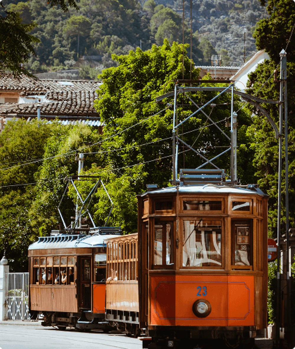 El emblemático tranvía vintage de madera atraviesa la histórica ciudad de Sóller, rodeada de exuberante vegetación y vistas a las montañas.